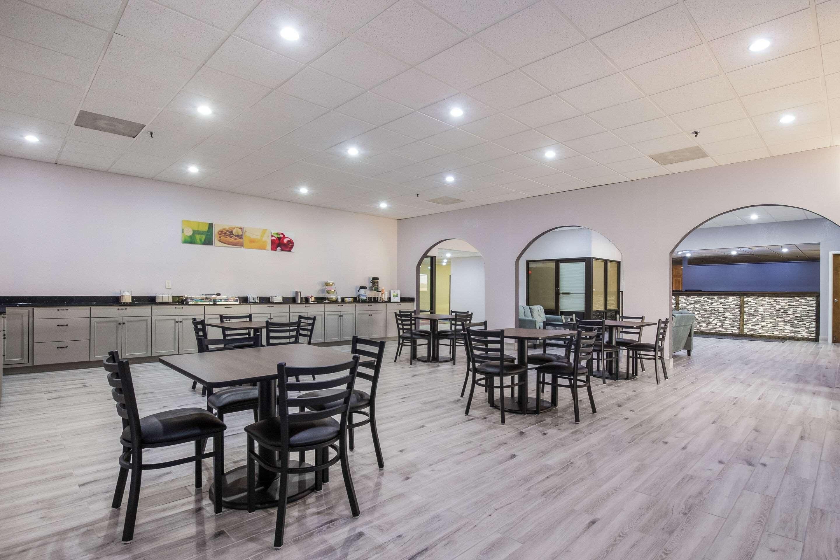Uma sala de jantar ampla com várias mesas e cadeiras, área de cozinha com armários, e vista através de uma janela em arco.