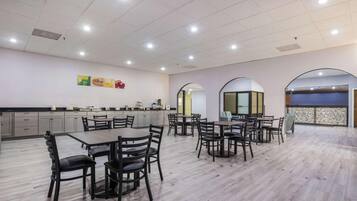 Uma sala de jantar ampla com várias mesas e cadeiras, área de cozinha com armários, e vista através de uma janela em arco.