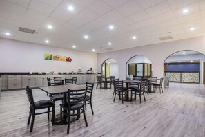 A spacious dining area with multiple tables and chairs, a kitchen area with cabinets, and a view through an arched window.