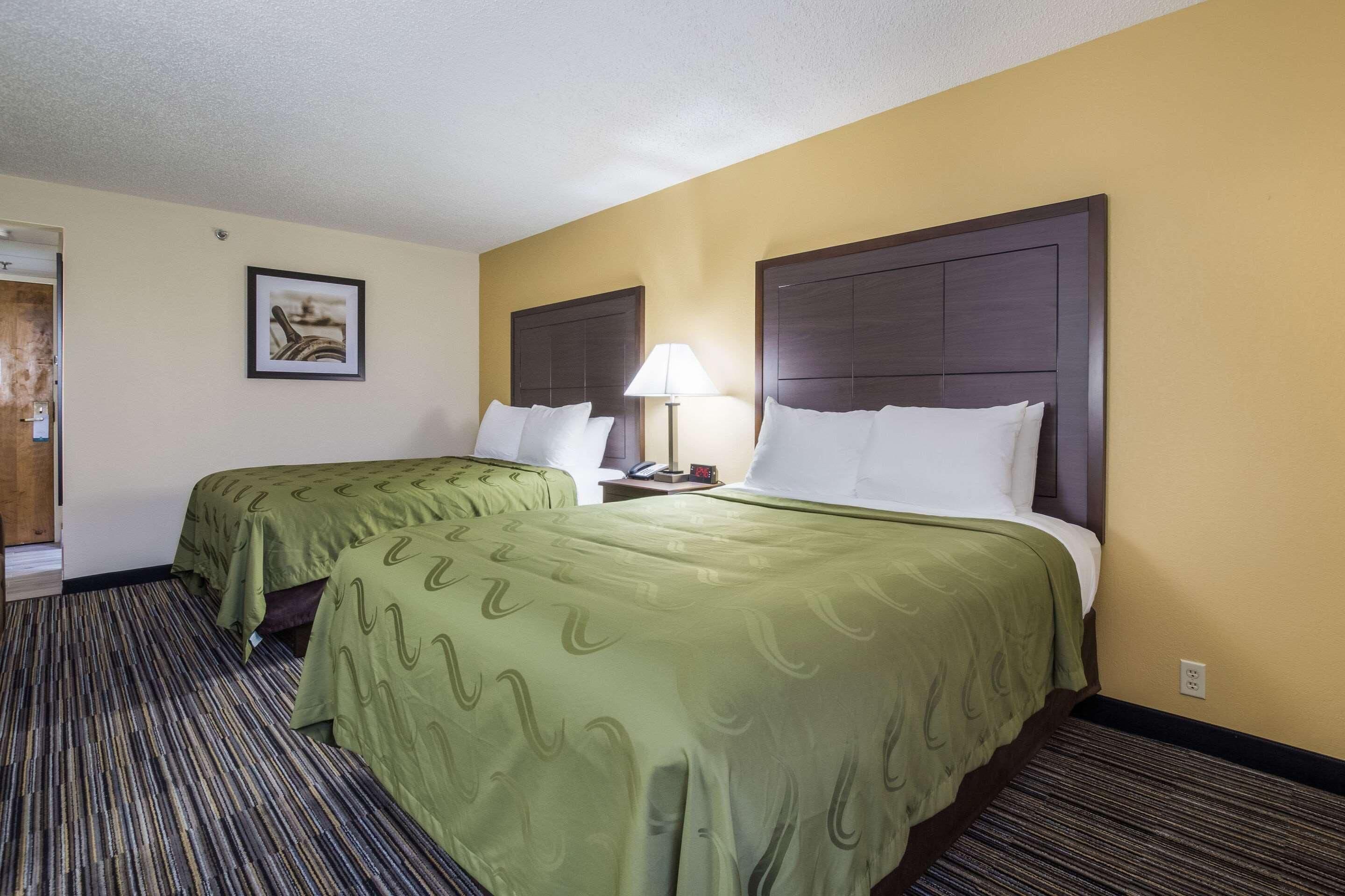 A hotel room with two beds, a carpeted floor, a lamp, and a framed picture on the wall.