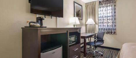 Una habitación de hotel con televisión de pantalla plana, mini refrigerador, escritorio con lámpara, una silla y una cama.