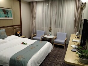A hotel room with a bed, two armchairs, a small table, a desk with a computer, and a potted plant.