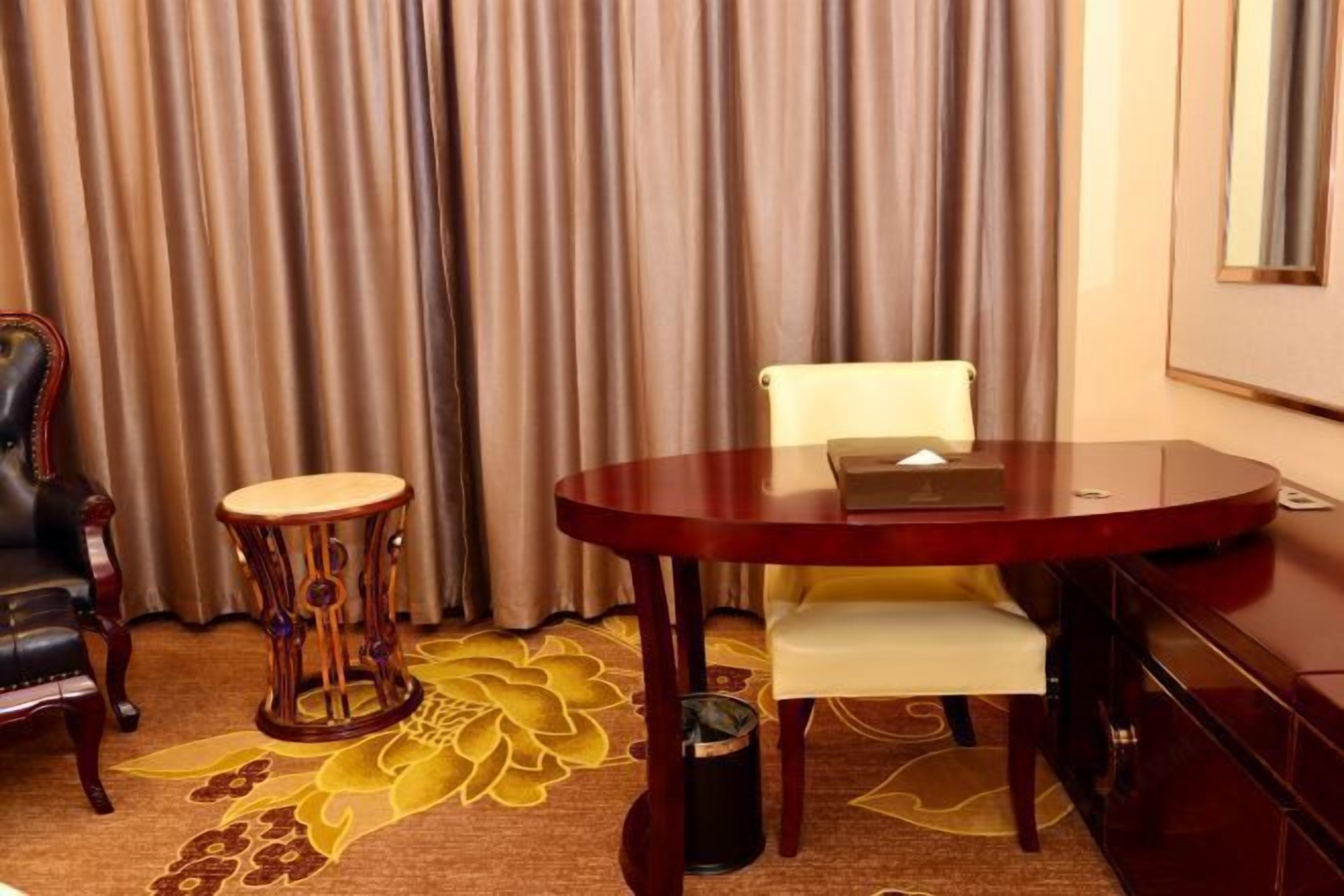 A hotel room with a round wooden table, a white cushioned chair, a decorative wooden stool, and a large floral carpet.
