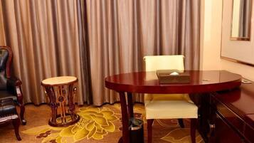 A hotel room with a round wooden table, a white cushioned chair, a decorative wooden stool, and a large floral carpet.