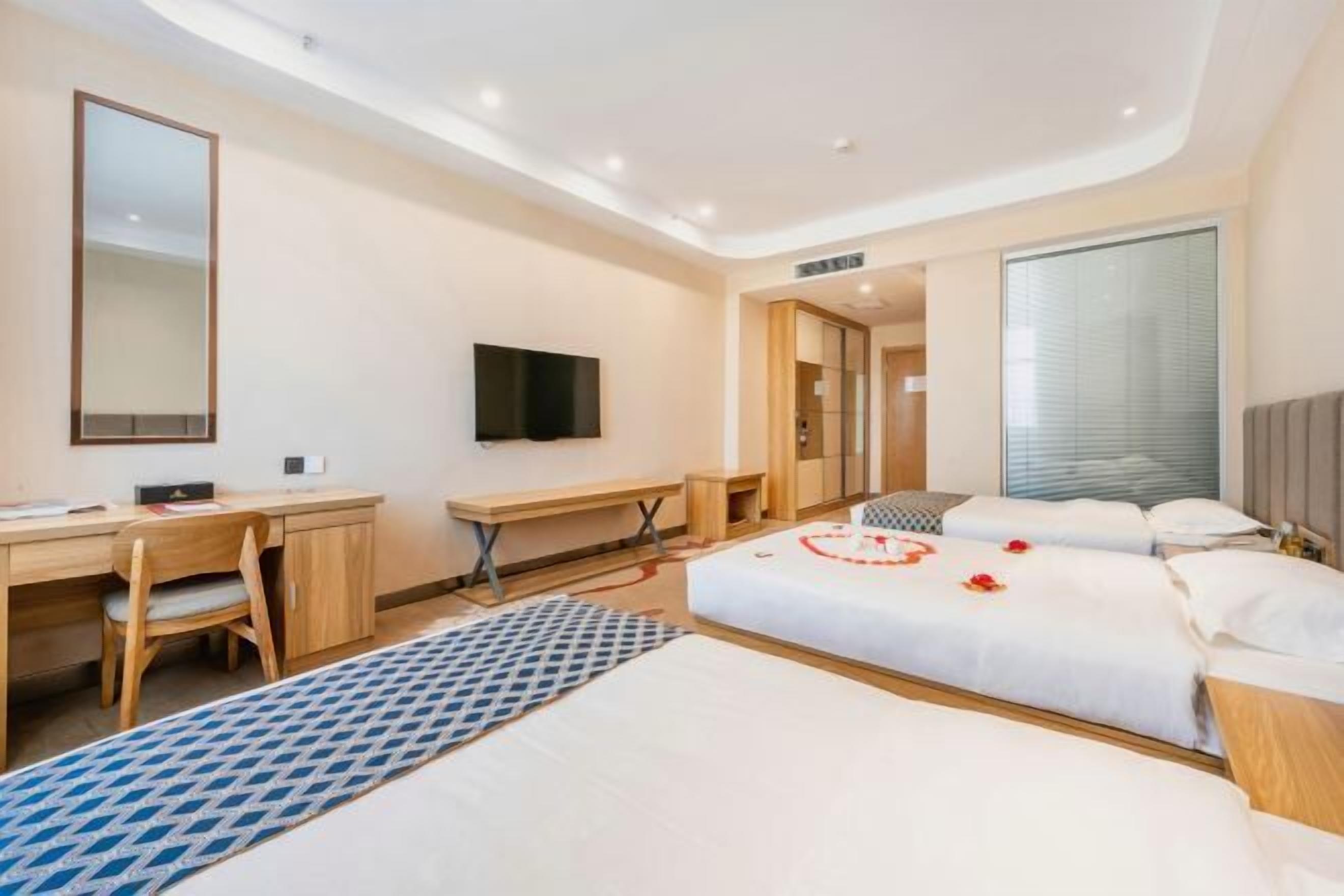 A hotel room with a desk, a bed with rose petals, a TV, and a large window.