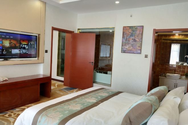 A hotel room with a bed, a TV mounted on the wall, a wooden desk, and a bathroom visible through an open door.