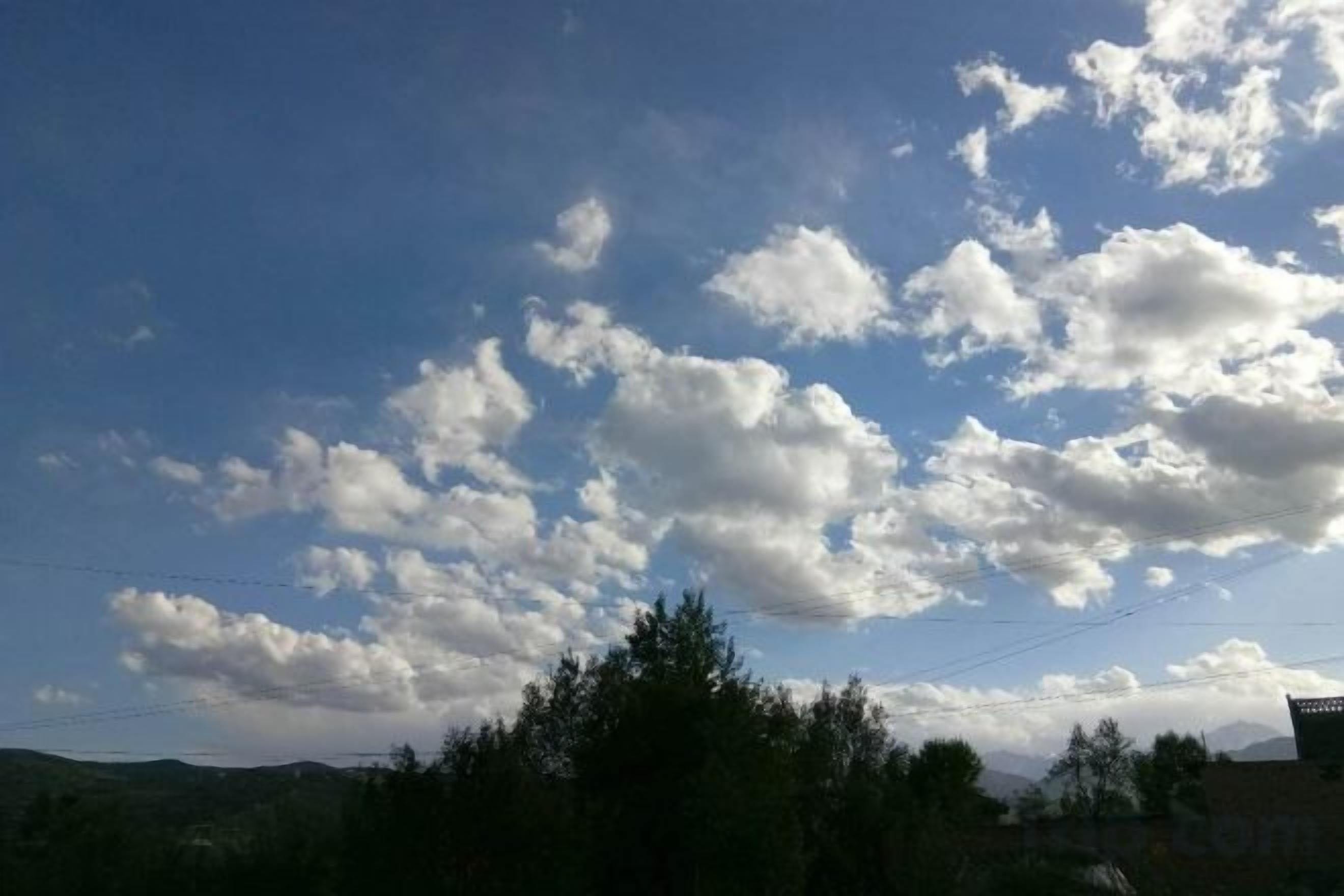 A clear sky with scattered clouds and a line of trees in the foreground.
