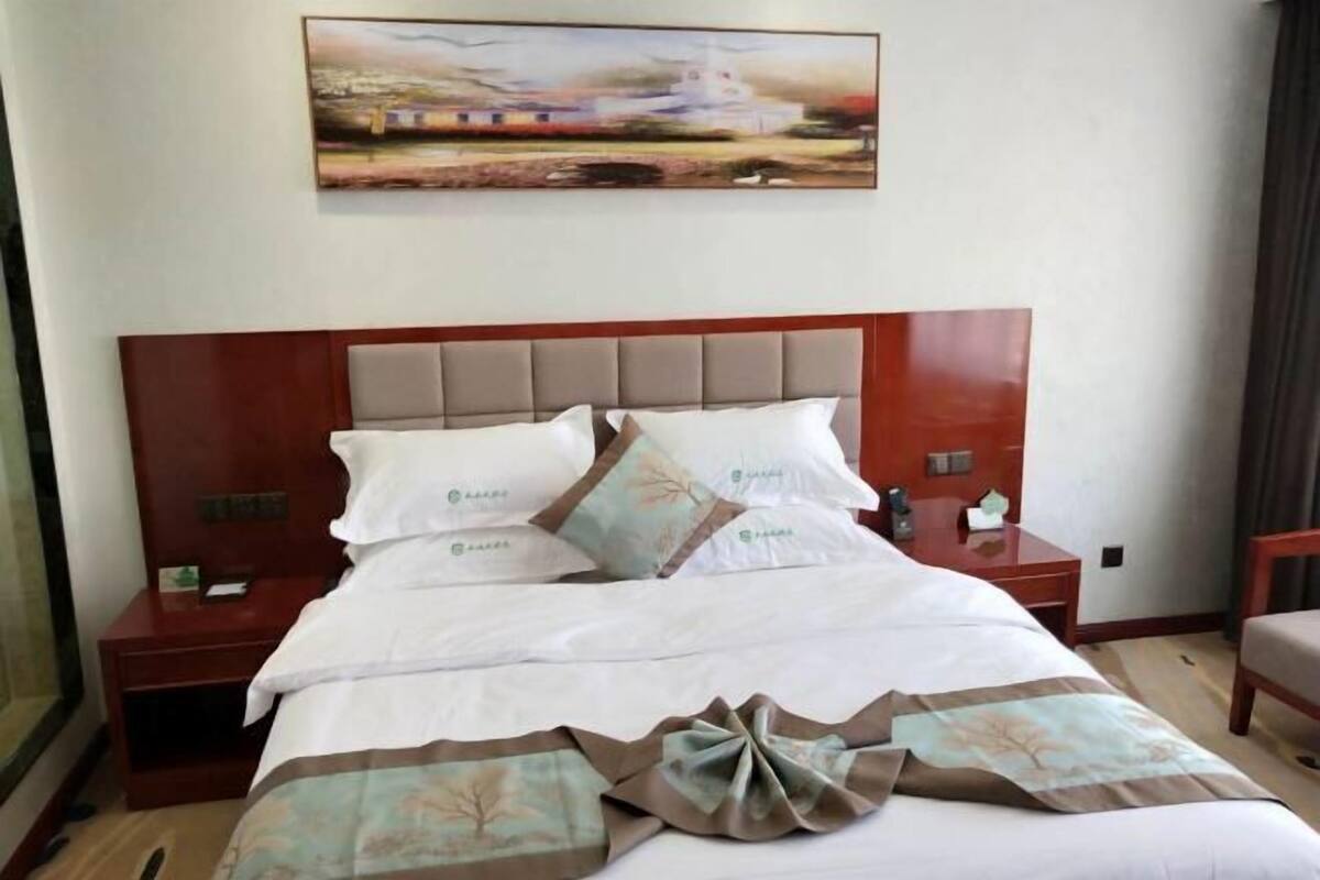 Una cama bien hecha con ropa blanca y un adorno decorativo, una mesita de noche de madera con una lámpara, y un cuadro sobre la cama.