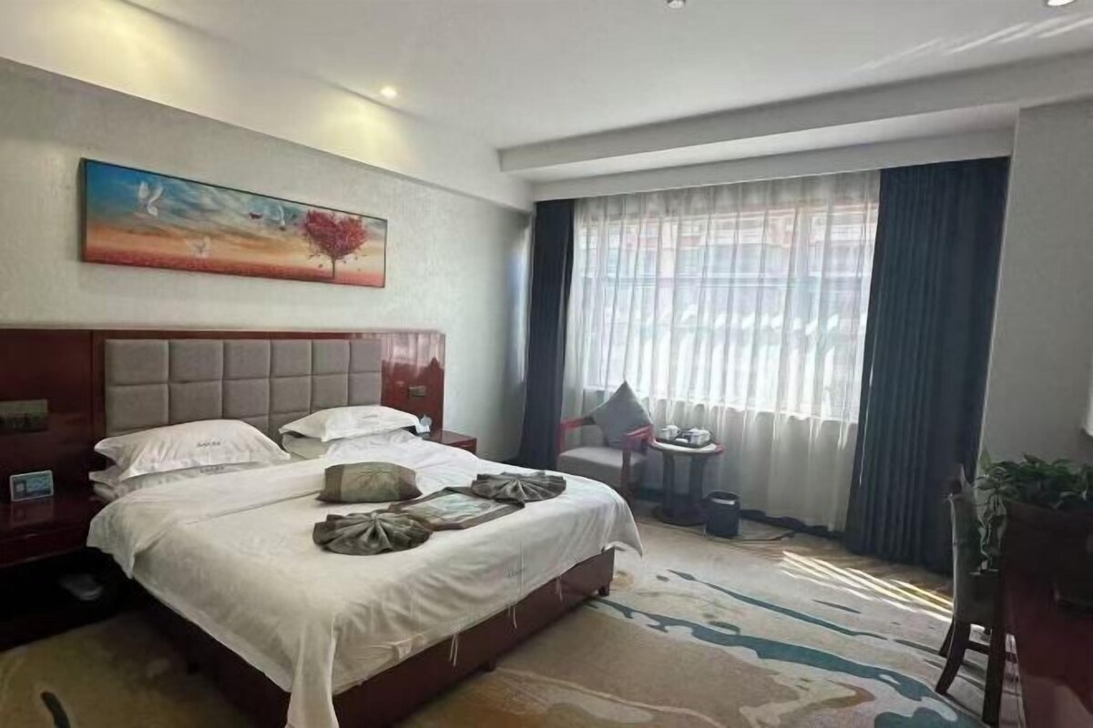 A hotel room with a large bed, a desk, a chair, a window with curtains, and a painting on the wall.