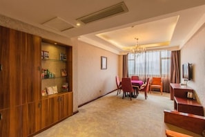 A hotel room with a wooden wardrobe, a dining table with chairs, a chandelier, and a television on the wall.