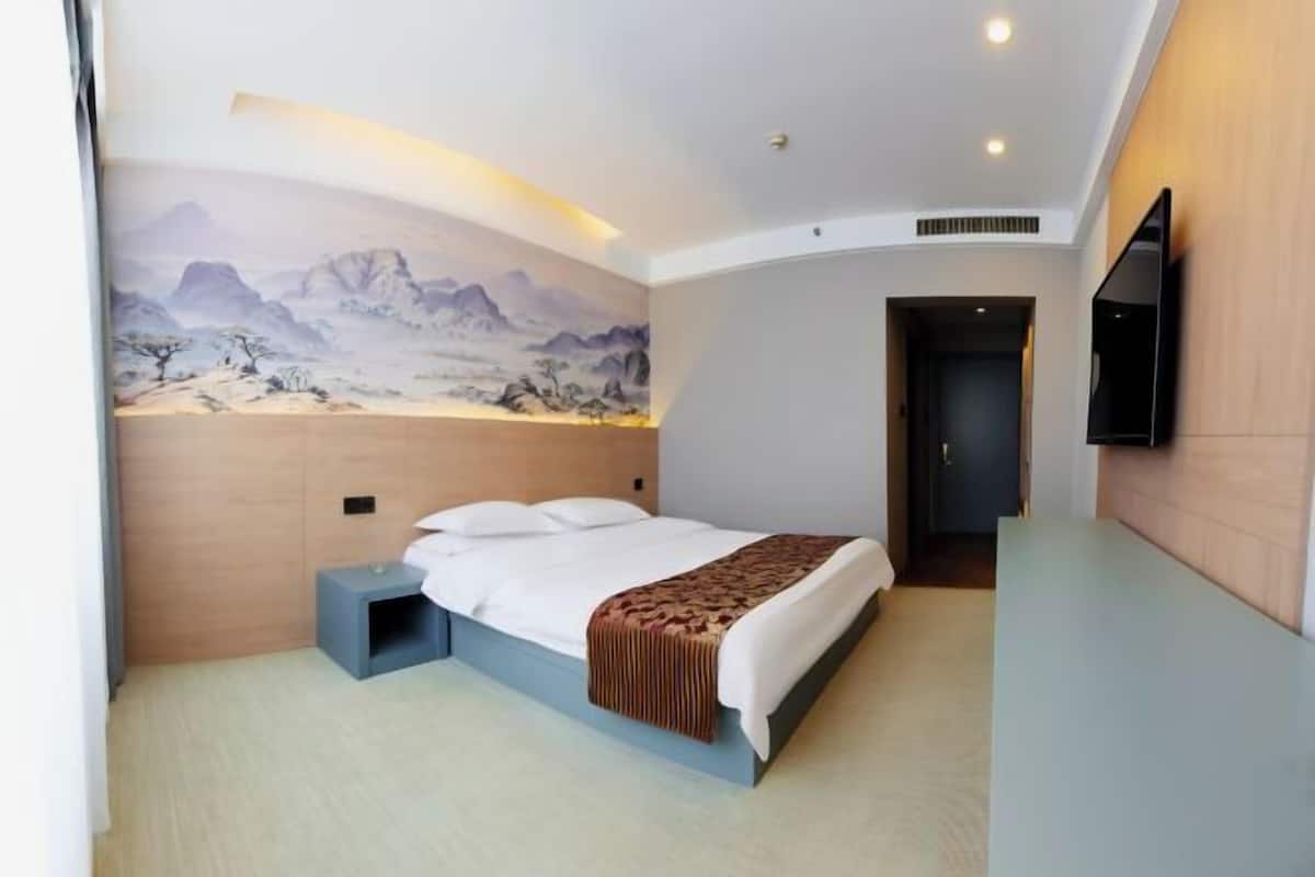 Una habitación de hotel moderna con una cama, una mesita de noche pequeña, un televisor montado en la pared y un cuadro de montañas.