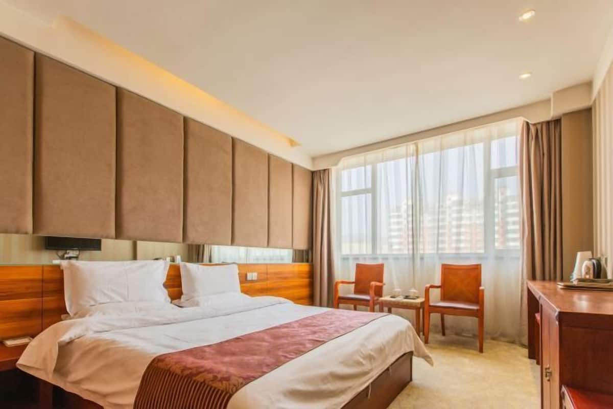 Habitación de hotel con una cama grande, cabecera de madera, un escritorio con silla y un ventanal con cortinas.