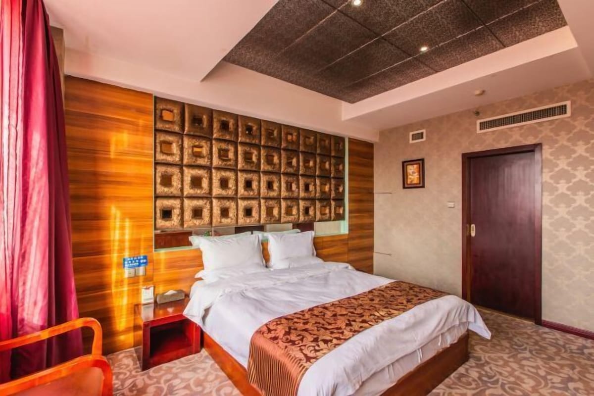 A hotel room with a bed, a nightstand, a red chair, a wooden headboard with square cutouts, a patterned carpet, and a dark door.
