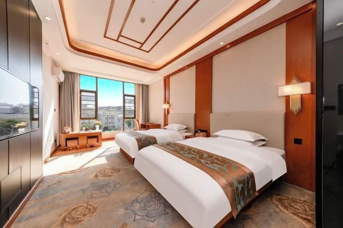 A hotel room with two beds, a desk, a TV, and a large window with a city view.