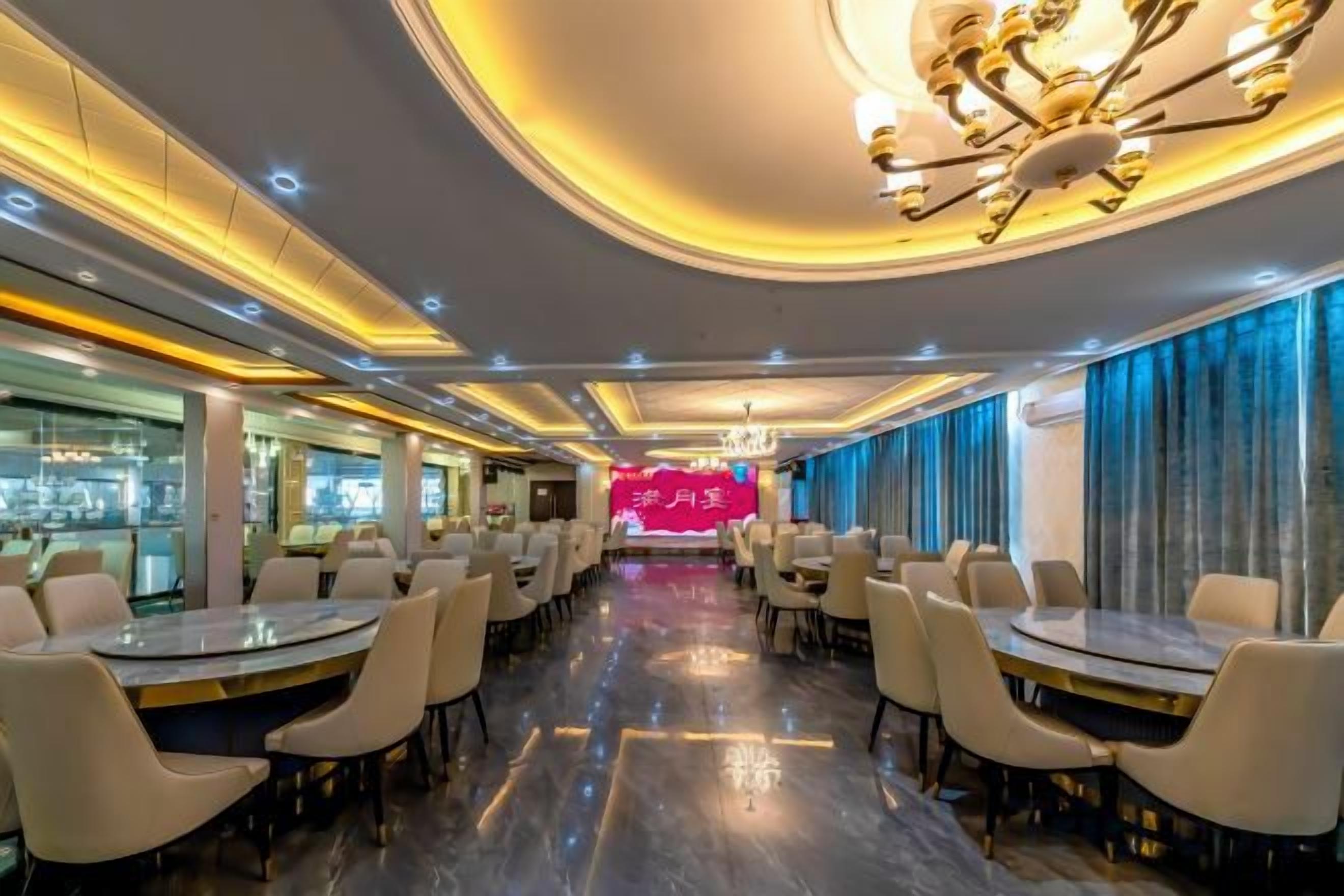 A hotel dining area with round tables and chairs, a bar on the left, and a stage with a red backdrop in the center.