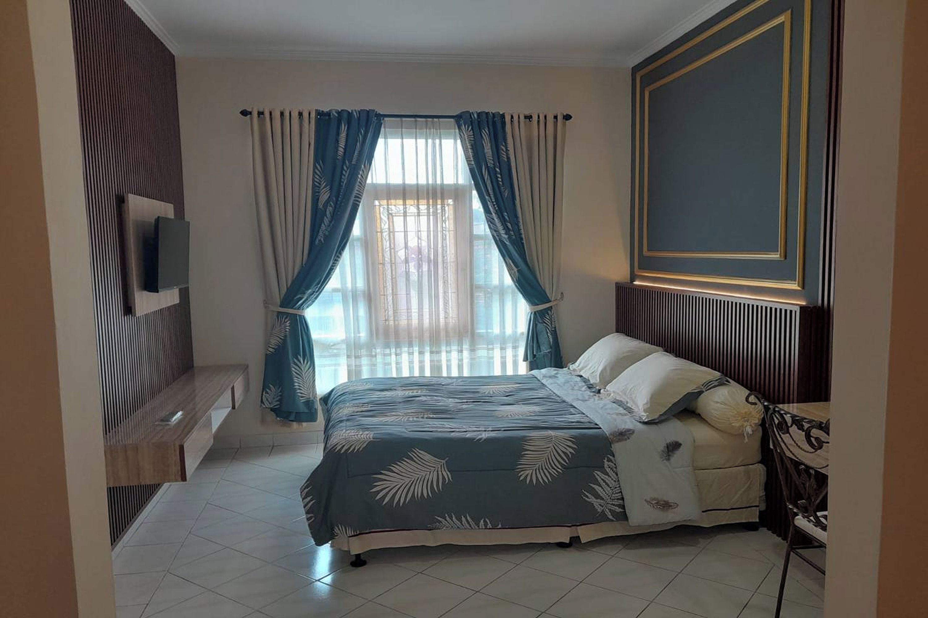 A bedroom with a bed, a TV mounted on the wall, a window with curtains, and a wooden bench.