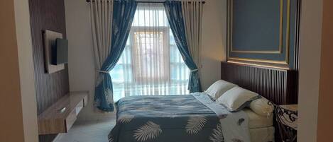 A bedroom with a bed, a TV mounted on the wall, a window with curtains, and a wooden bench.