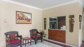 A reception area with a wooden reception desk, two wooden chairs with red cushions, a framed painting of a street scene, and a decorative wall piece.
