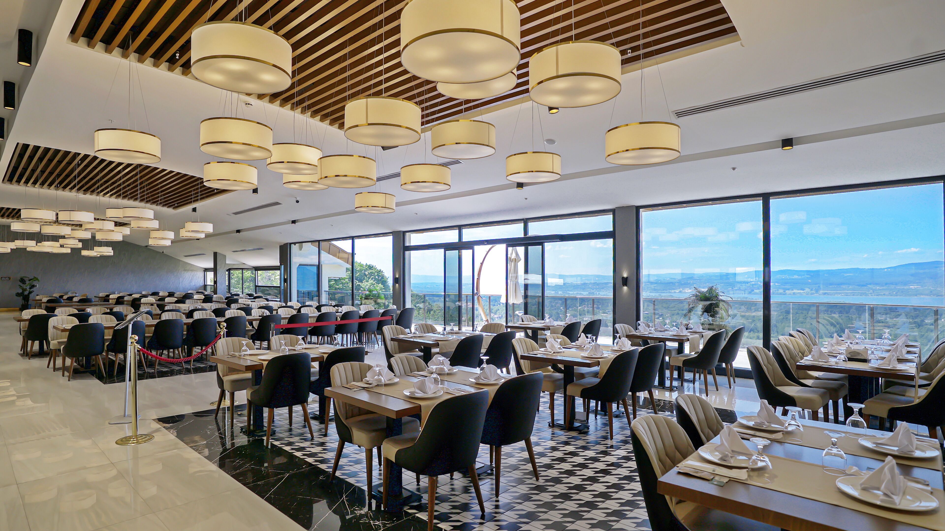 Un restaurante espacioso con techos altos, candelabros modernos y grandes ventanales que ofrecen una vista panorámica del paisaje.