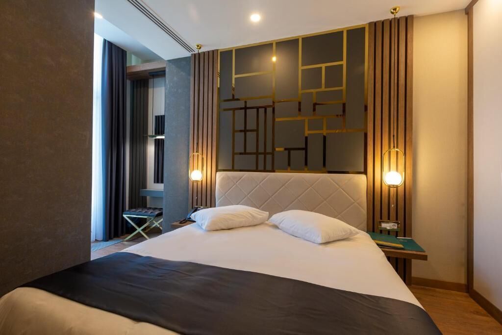 Una habitación de hotel moderna con una cama grande, cabecera con diseños geométricos, lámparas de noche y una mesita con un libro.