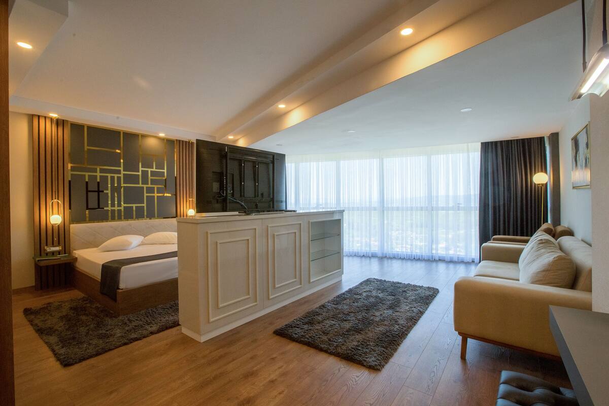 Una habitación de hotel moderna con cama, sofá, zona de cocina y un ventanal con cortinas.