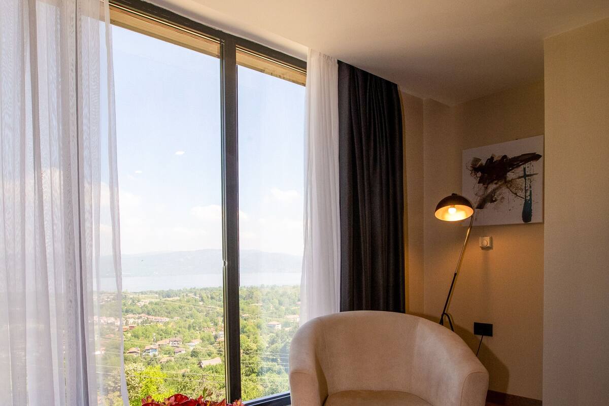 Una habitación con un ventanal grande, una silla, una mesa con flores, una lámpara de pie y un cuadro en la pared.