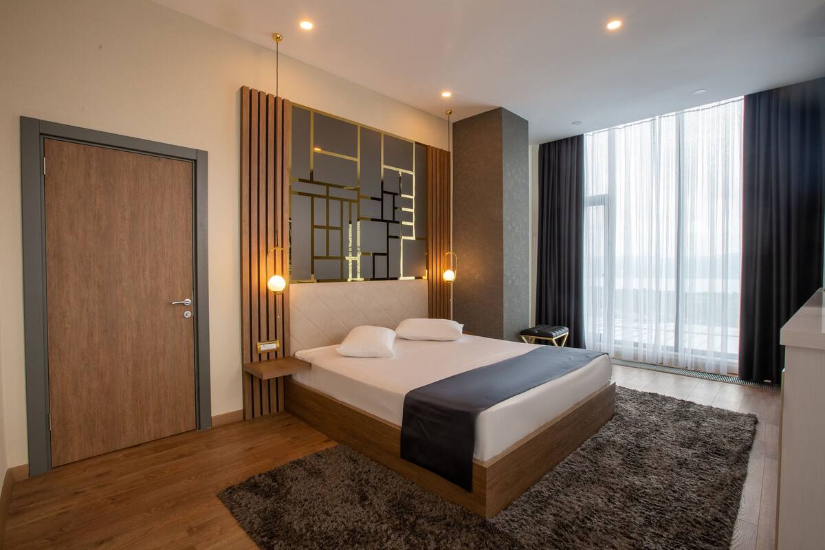Un dormitorio moderno con una cama grande, una puerta de madera, un diseño geométrico en la pared y un amplio ventanal con cortinas.