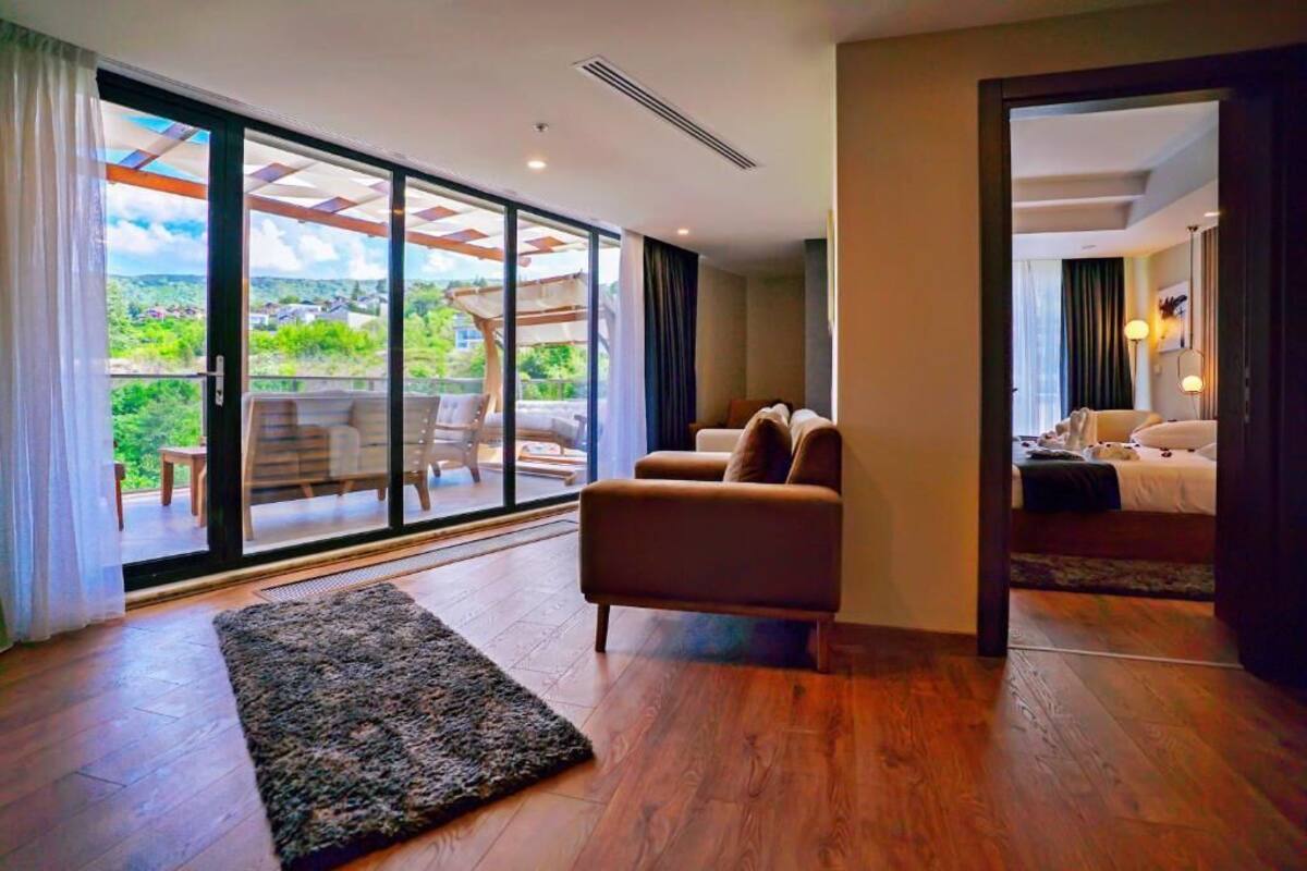 Una habitación de hotel moderna con un amplio ventanal corredera que da a un balcón, un sofá, una cama y una pequeña alfombra sobre el suelo de madera.
