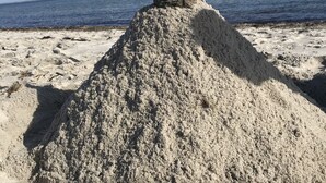 Une structure de sable surmontée d’une pile de pierres équilibrées, située sur une plage avec vue sur l’océan.