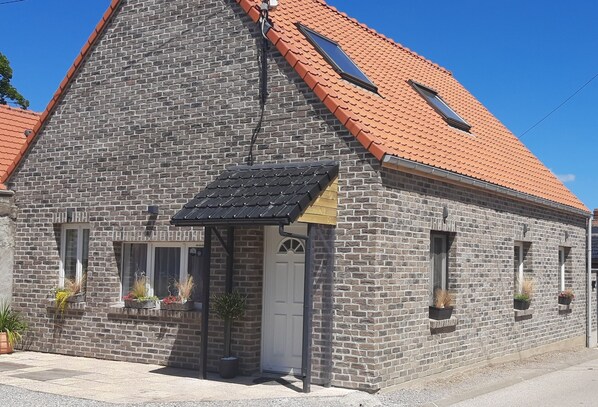 Ein Steinhaus mit rotem Ziegeldach, ein kleiner Anbau mit schwarzem Markisenstoff und Kübelpflanzen in Fensterkästen.