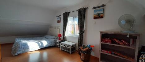 Un dormitorio con cama, una mesita de noche con lámpara, una cesta con juguetes, una estantería y una ventana con cortinas.
