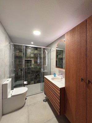 A modern bathroom with a glass-enclosed shower, a white toilet, a wooden vanity, and a large mirror.
