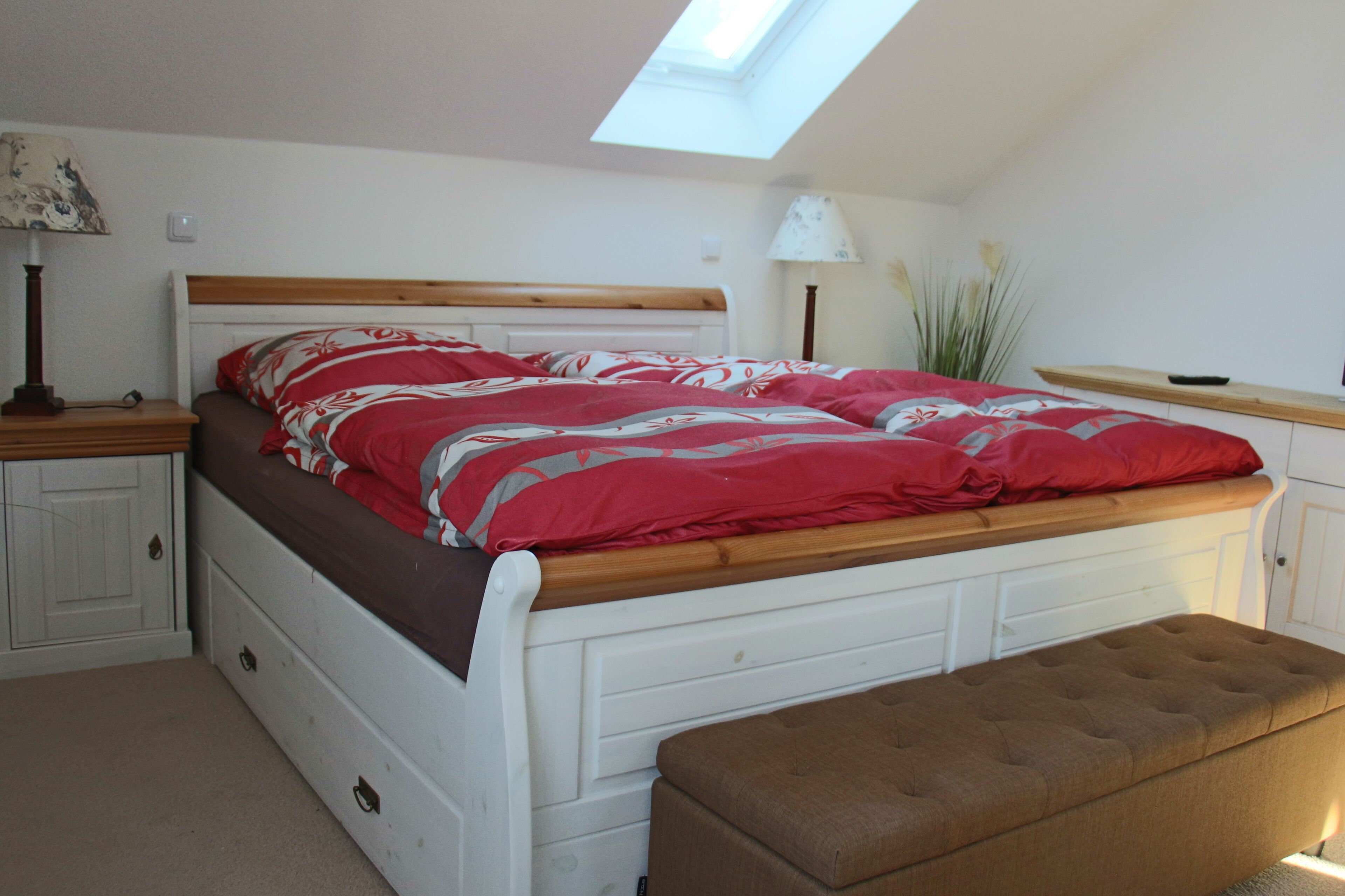 Un dormitorio con tragaluz, cama con colcha de diseño rojo y blanco, cabecero de madera, mesita de noche con lámpara y un banco al pie de la cama.