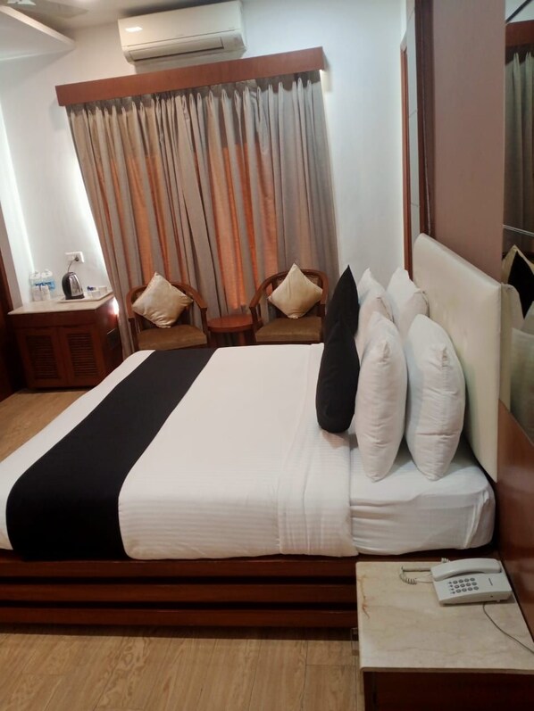 A hotel room with a bed, a bedside table with a phone, a wooden headboard, a window with curtains, and a wall-mounted air conditioner.