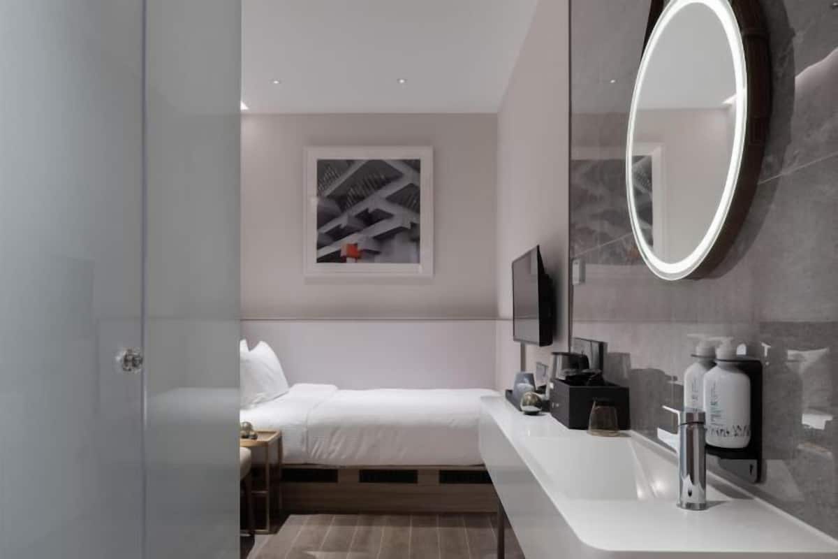 A modern hotel room with a bed, a large mirror, a TV, and a bathroom with a sink and toiletries.