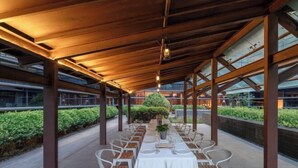 An outdoor dining area with a long table set for a meal, surrounded by white chairs, under a wooden pergola with glass panels. - MeiChen Hotel( within the terminal building of Beijing Daxing International Airport) (Beijing)