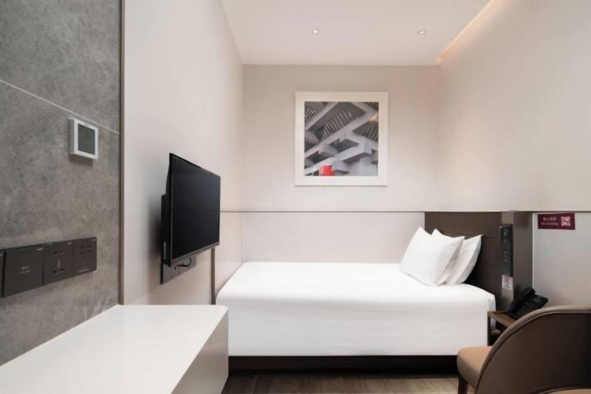 A modern hotel room with a single bed, a flat-screen TV mounted on the wall, a small window with a cityscape view, and a desk with a chair.
