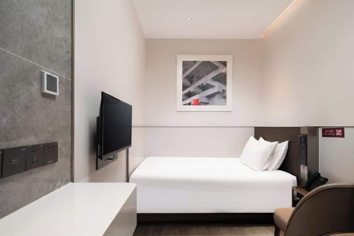 A modern hotel room with a bed, a flat-screen TV, a small window with a cityscape view, a bedside table, and a chair.