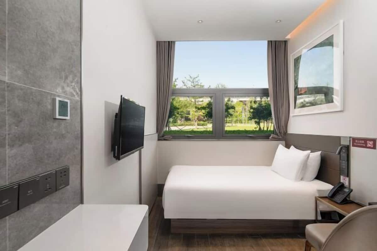 A hotel room with a bed, a TV mounted on the wall, a window with a view of greenery, and a bedside table with a phone.