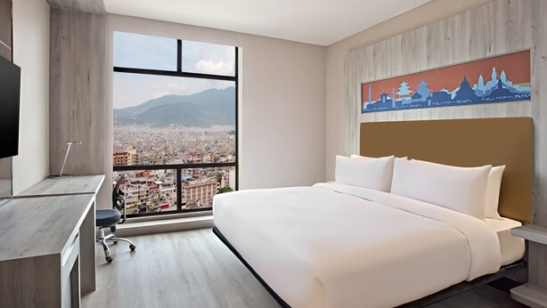 A hotel room with a large bed, a desk with a chair, a TV, and a view of a cityscape through a large window. - Aloft Kathmandu Thamel (Kathmandu)