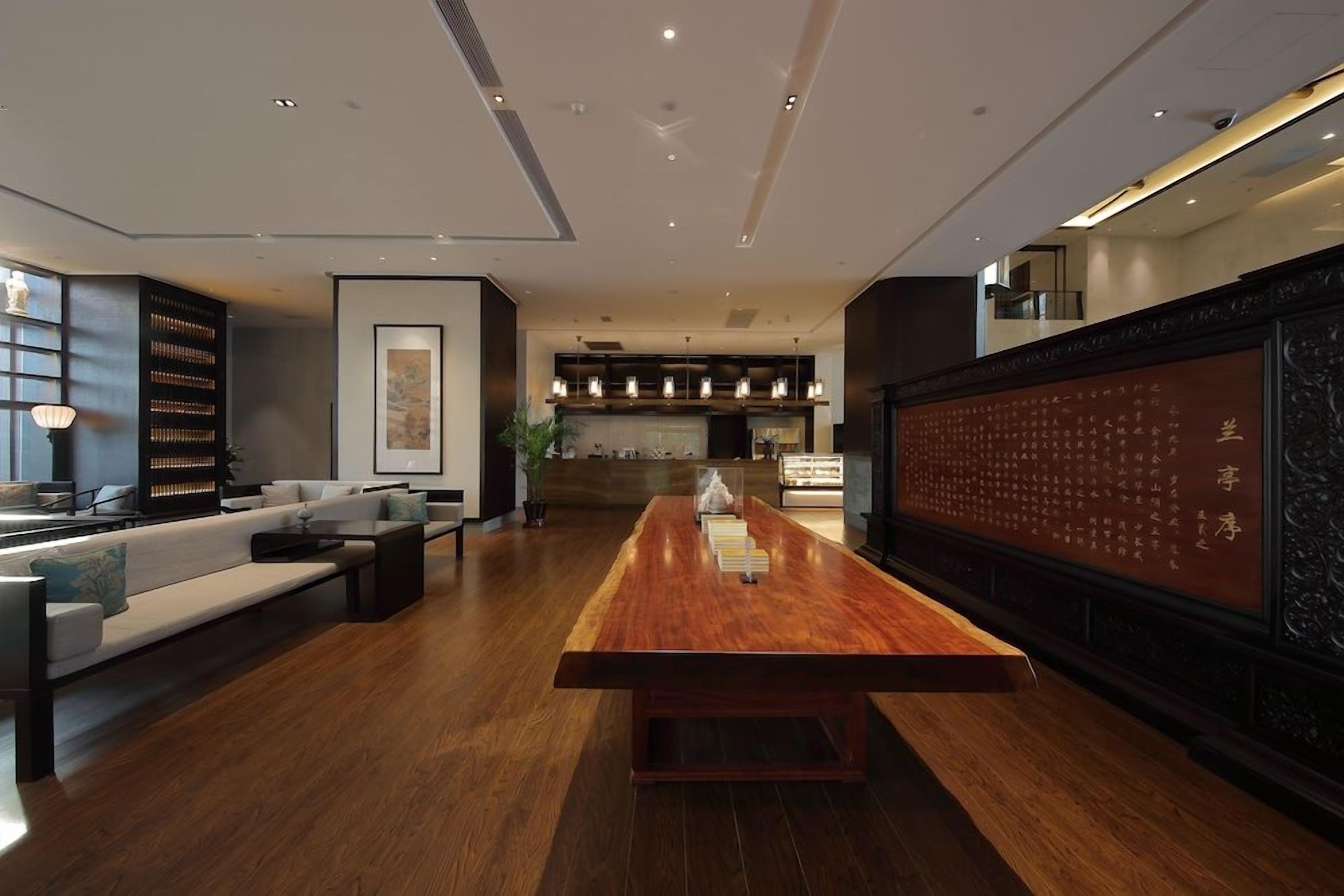 A modern hotel lobby with a long wooden reception desk, seating area, and a decorative wall with calligraphy.