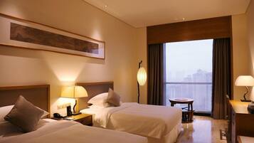 Quarto de hotel com duas camas, uma mesinha, uma luminária, um quadro na parede e vista da cidade pela janela.