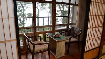 Una habitación tradicional japonesa con suelo de tatami, una mesa de madera baja, dos sillas y ventanas amplias con vistas a árboles.