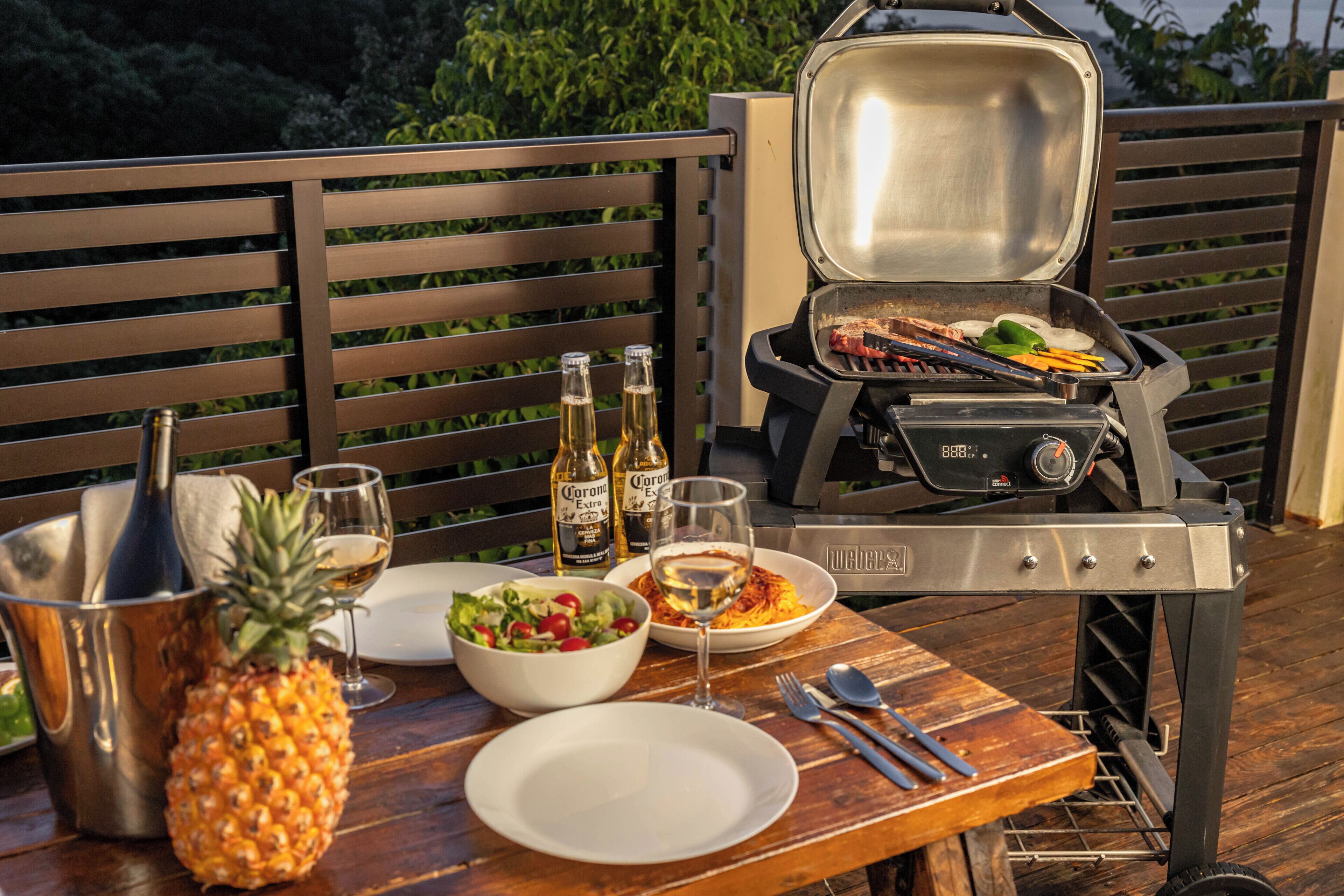 Un barbecue avec de la nourriture, un ananas, des bouteilles et une salade, le tout posé sur une table en bois.
