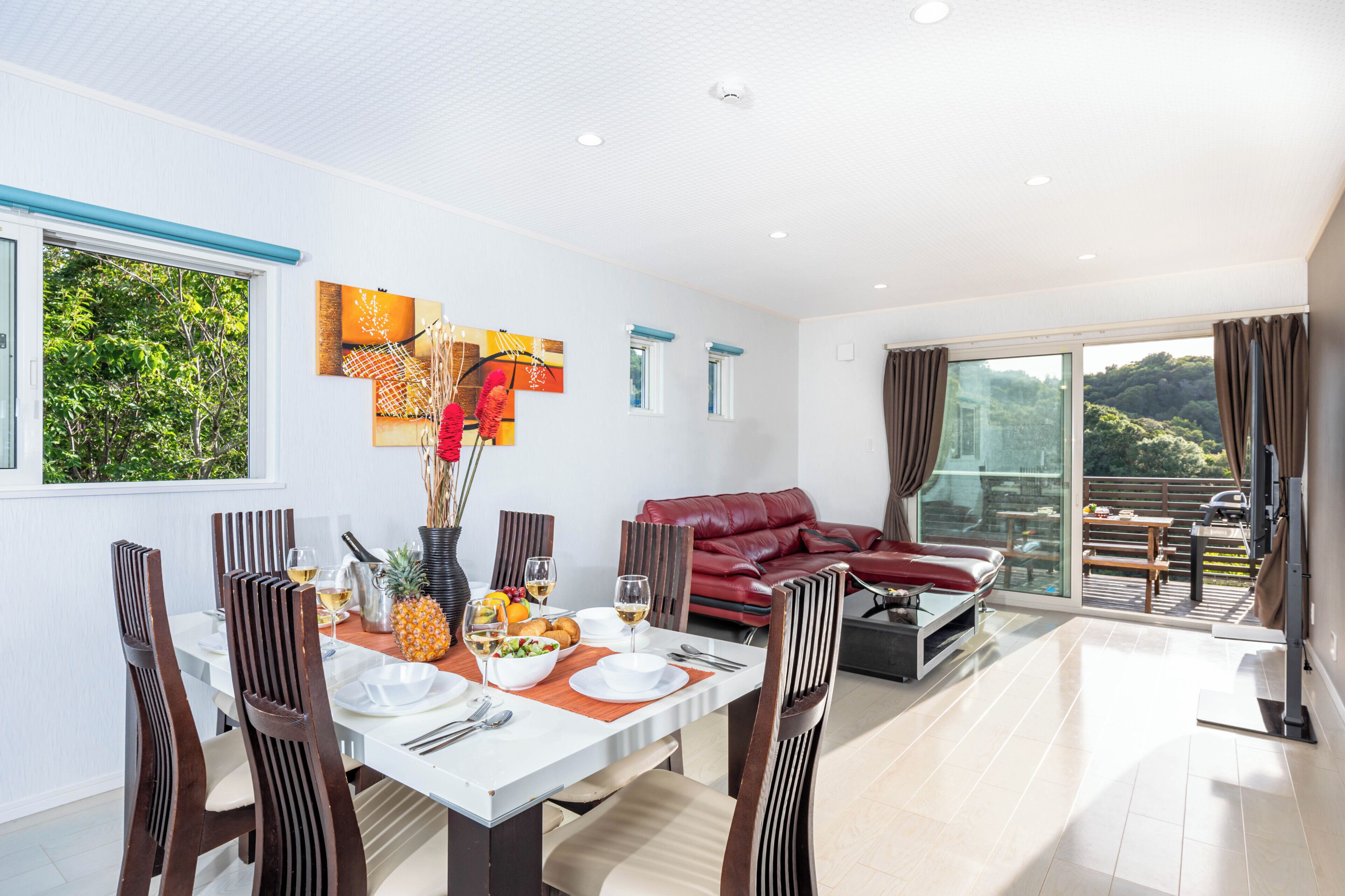 Uma área de refeições com mesa posta para quatro pessoas, um sofá de couro vermelho e uma varanda com mobiliário de exterior.