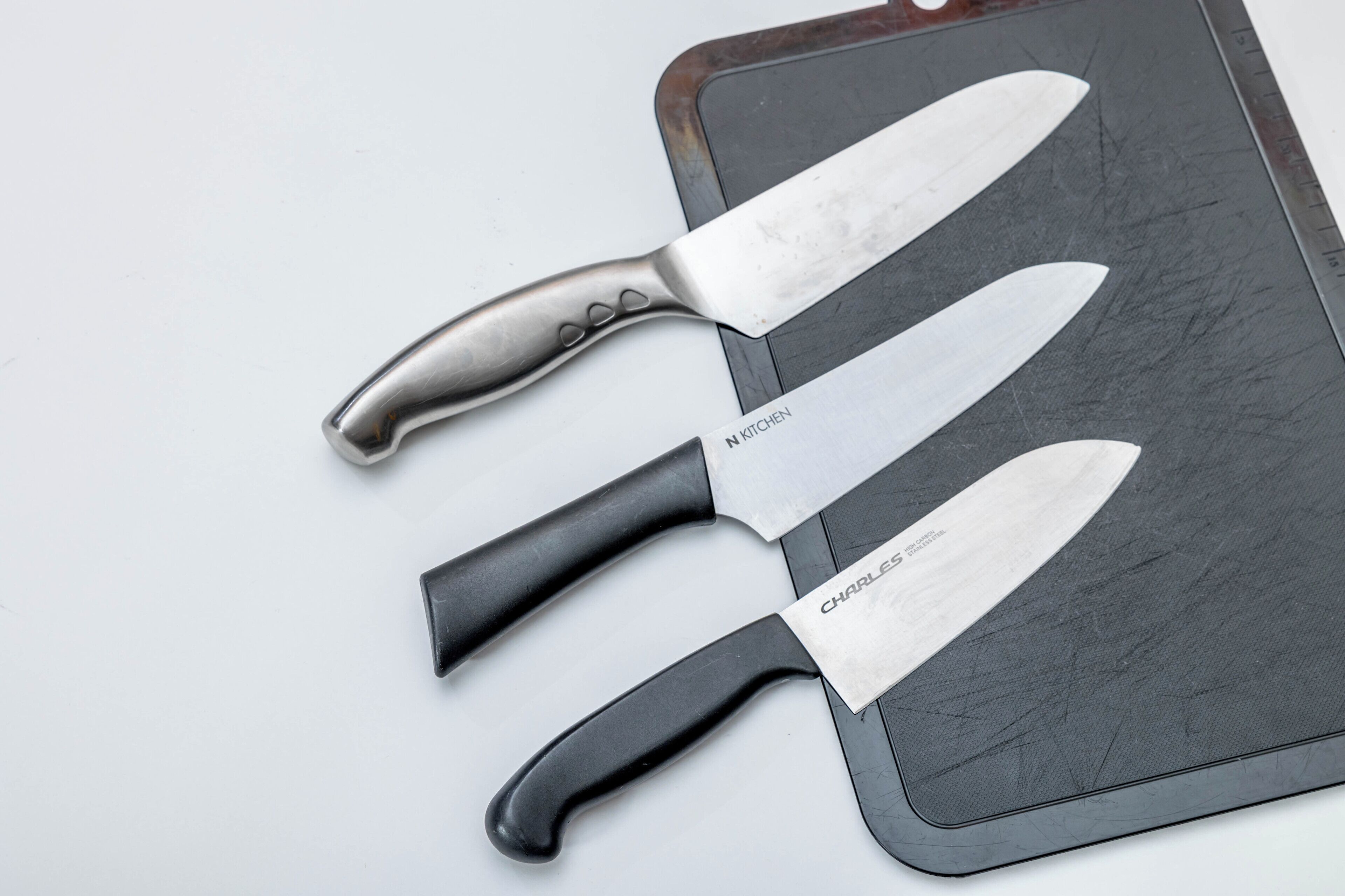 Trois couteaux de chef à manche noir et une planche à découper noire, le tout posé sur une surface blanche.