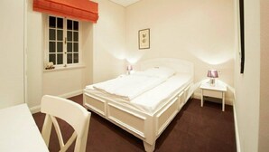 Ein Schlafzimmer mit Bett, Nachttischen, einem Fenster mit Vorhängen, einem Sessel und einem kleinen Tisch.