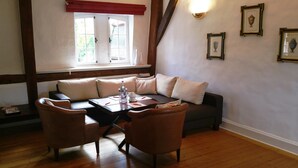 Ein Zimmer mit einer Couch, zwei Sesseln, einem Tisch mit einer Flasche und Gläsern, einem Fenster mit Rollläden und gerahmten Bildern an der Wand.