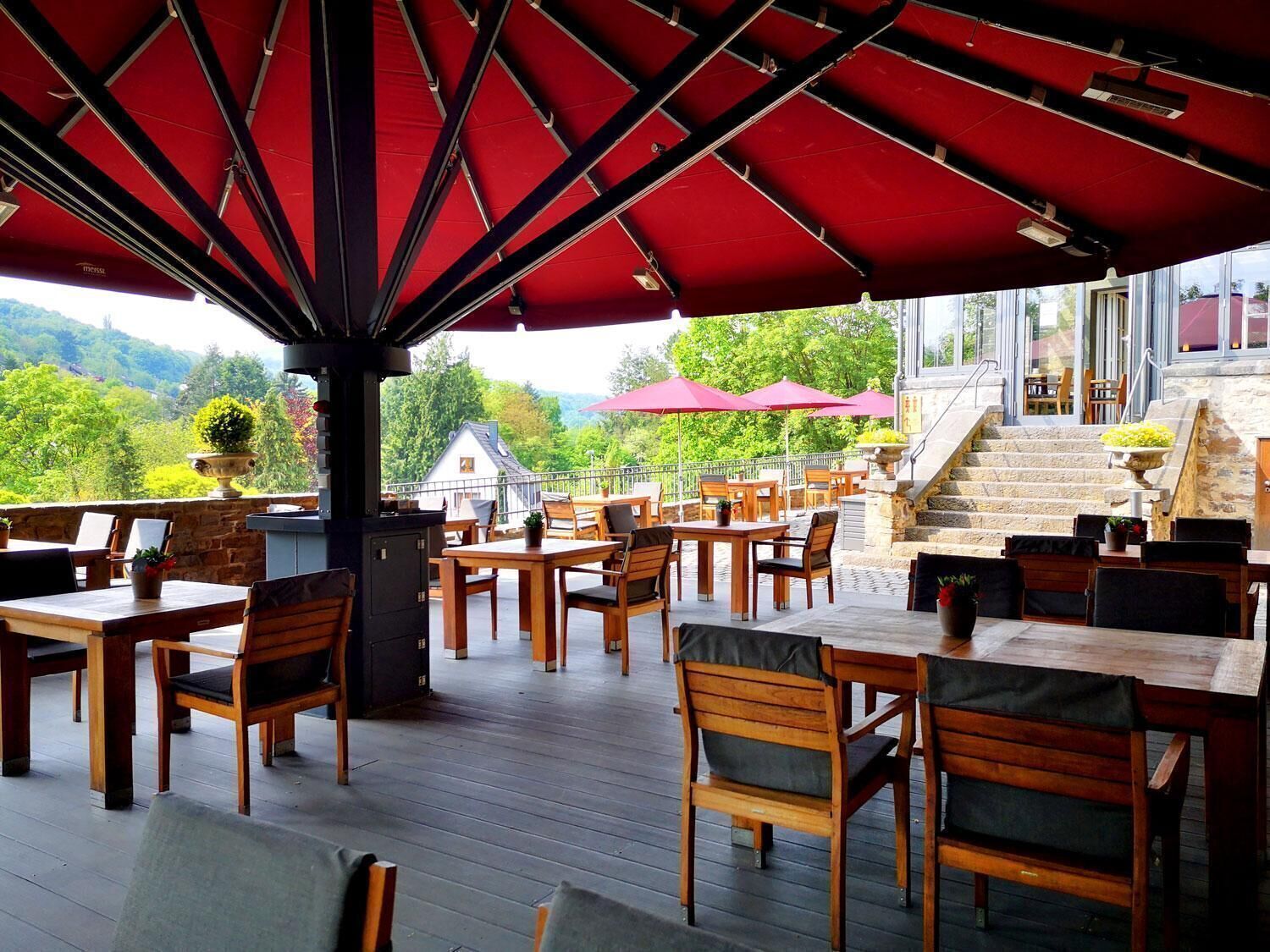 An outdoor patio with wooden tables and chairs, a red umbrella, and a view of a house and greenery.