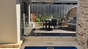 Une piscine avec une terrasse carrelée, un espace patio avec une table et des chaises, et une clôture en bois en arrière-plan.